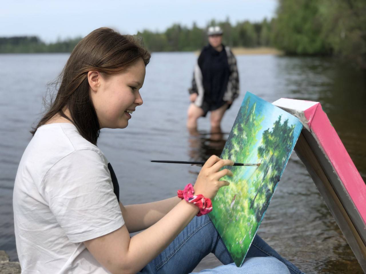 Opiskelija maalaa rantaviivassa istuen, toinen opiskelija kahlaa taustalla
