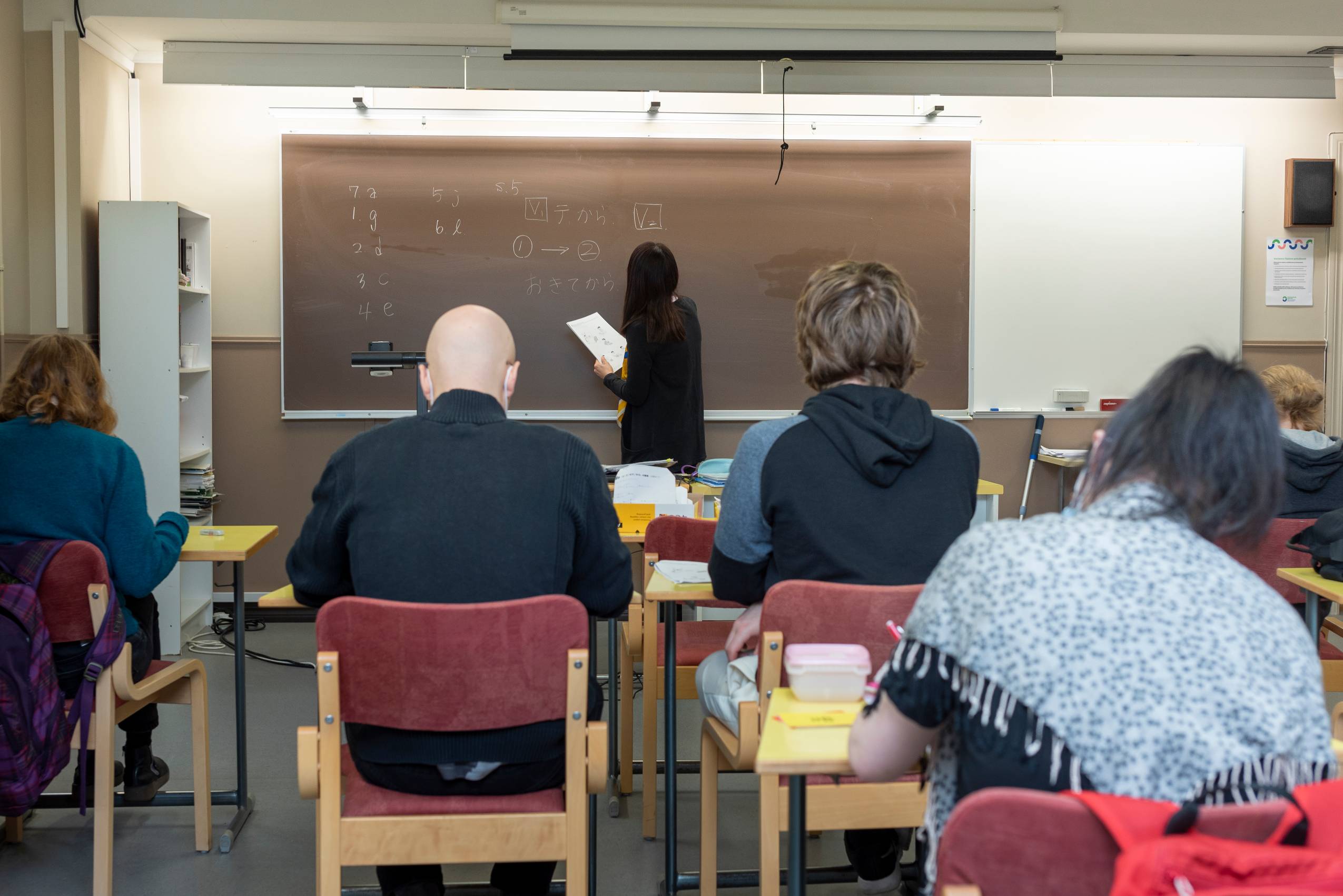 Japanin opettaja kirjoittaa taululle japanilaista tekstiä, kaksi opiskelijaa kuvassa